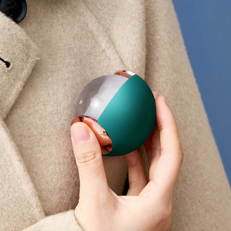 Hand holding green and silver reusable lint remover ball on beige coat with blue background