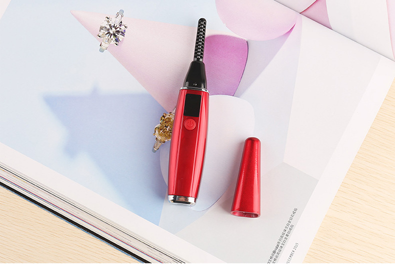 Red heated eyelash curler with temperature display on a vanity with makeup items